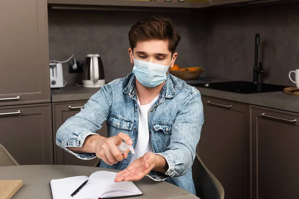 Homme en masque médical utilisant un désinfectant pour les mains près d'un ordinateur portable sur une table de cuisine — Photo de stock
