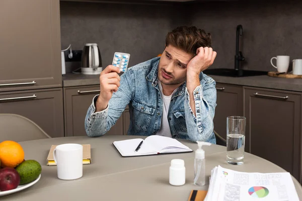 Focus selettivo di uomo triste che tiene blister con pillole vicino a carte, notebook e disinfettante per le mani sul tavolo della cucina — Foto stock