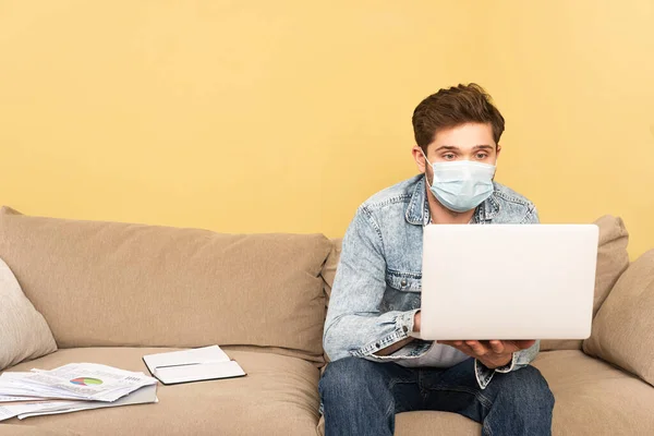 Freelancer en máscara médica usando laptop cerca de notebook y documentos en sofá - foto de stock