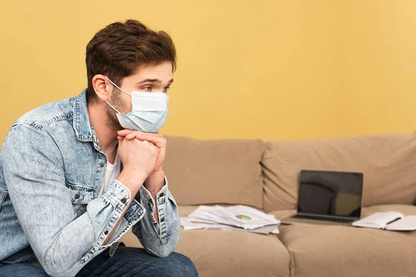 Focus selettivo del libero professionista in maschera medica seduto vicino al computer portatile e carte sul divano — Foto stock