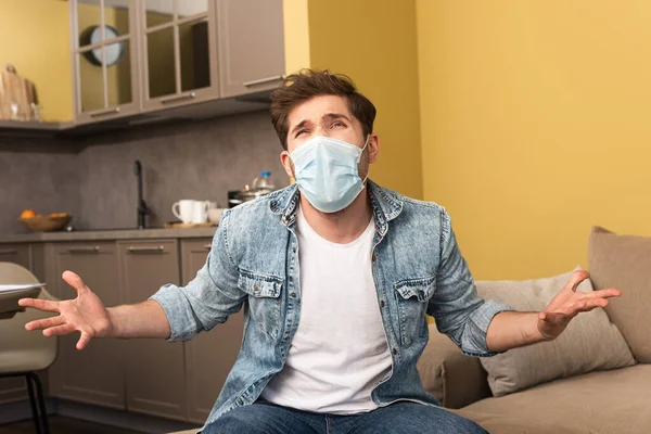 Недовольный мужчина в медицинской маске сидит дома на диване — стоковое фото