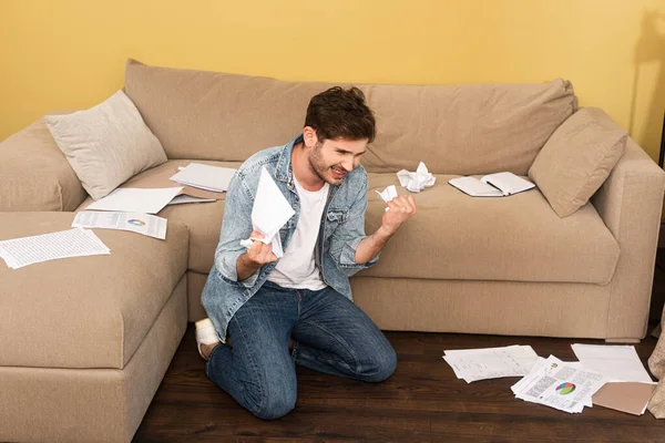Агресивний чоловік тримає згорнутий папір біля документів на дивані і підлозі у вітальні — стокове фото