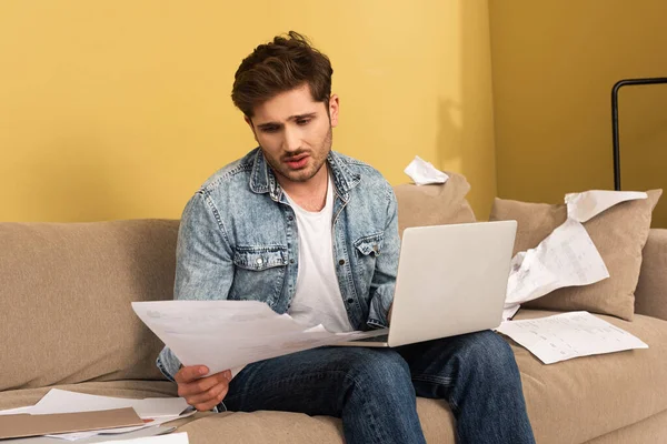 Грустный человек работает с бумагами и ноутбуком на диване дома — стоковое фото