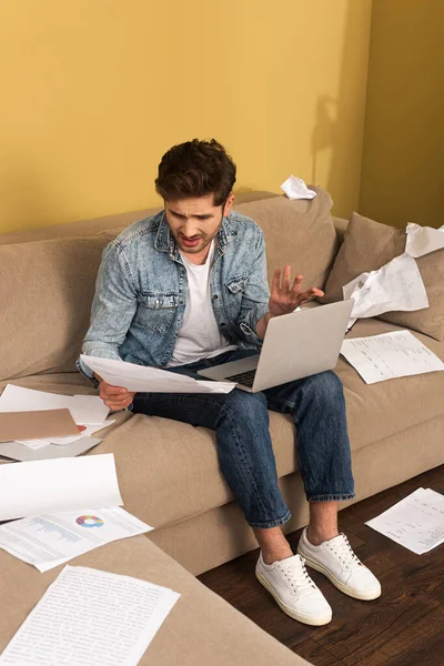 Нещасний чоловік тримає документ під час роботи з ноутбуком на дивані у вітальні — стокове фото