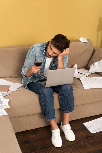 Freelancer pensativo segurando copo de vinho perto de laptop e papéis no sofá em casa — Fotografia de Stock