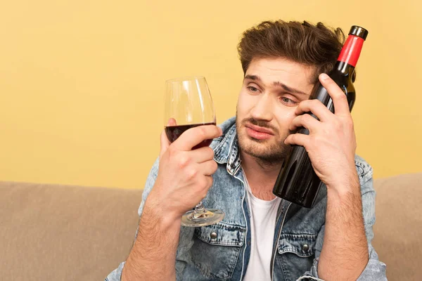 Upset man looking at glass of wine while holding bottle on sofa in living room — Stock Photo
