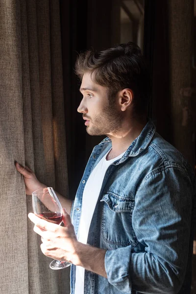 Seitenansicht eines gutaussehenden Mannes, der zu Hause ein Glas Wein in der Nähe des Fensters hält — Stockfoto