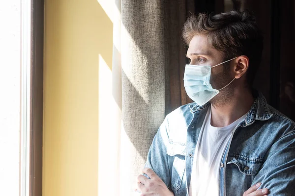 Man in medical mask with crossed arms looking at window in living room — Stock Photo