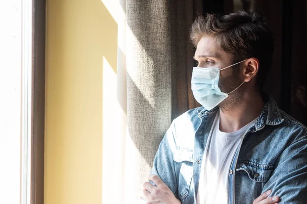 Человек в медицинской маске со скрещенными руками смотрит в окно в гостиной — стоковое фото