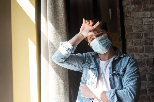 Man in medical mask with hand near forehead standing near window at home — Stock Photo