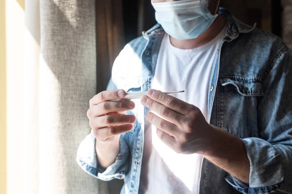 Vista recortada del hombre en máscara médica que sostiene el termómetro cerca de la ventana en casa - foto de stock