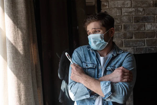 Больной человек в медицинской маске со скрещенными руками держит термометр возле окна в гостиной — стоковое фото