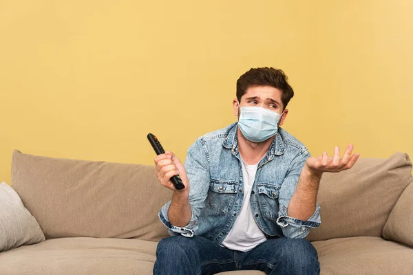 Hombre confuso con máscara médica sosteniendo mando a distancia en casa - foto de stock