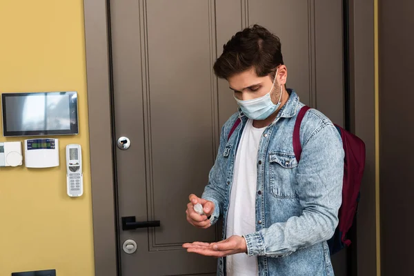 Jeune homme en masque médical utilisant un désinfectant pour les mains près de la porte à la maison — Photo de stock
