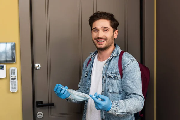 Hombre guapo en guantes de látex sonriendo a la cámara mientras que el agujero máscara médica en el pasillo - foto de stock