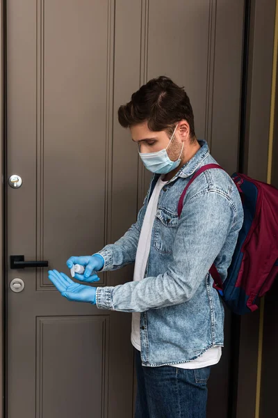 Vue latérale de l'homme en gants de latex et masque médical à l'aide d'un désinfectant pour les mains près de la porte à la maison — Photo de stock