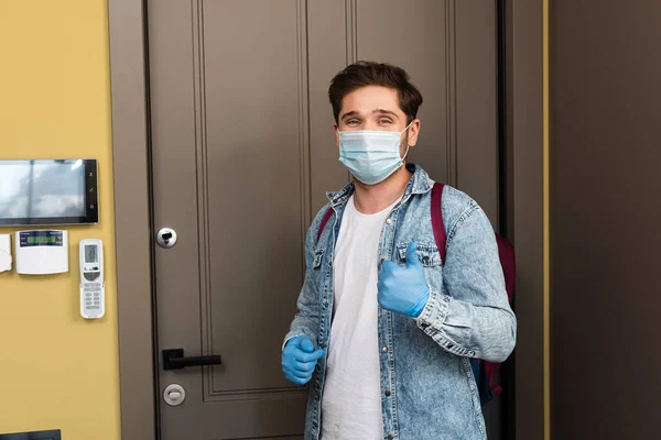 Hombre con máscara médica y guantes de látex mostrando como gesto cerca de la puerta en el pasillo - foto de stock