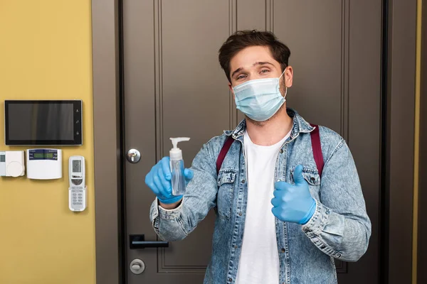 Hombre con máscara médica y guantes de látex mostrando el pulgar hacia arriba y sosteniendo el desinfectante de manos cerca de boor en casa - foto de stock