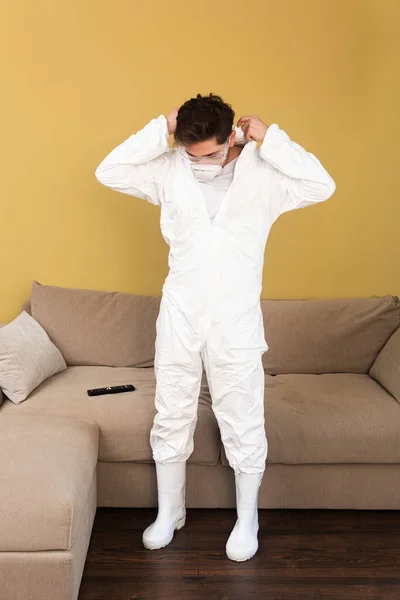 Uomo in maschera medica e occhiali di sicurezza indossando tuta hazmat in soggiorno — Foto stock