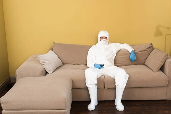 Mann in Schutzanzug und medizinischer Maske hält Fernbedienung auf Couch — Stockfoto