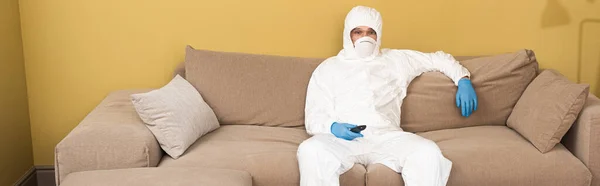 Panoramic shot of man in hazmat suit, medical mask and latex gloves holding remote controller on couch — Stock Photo