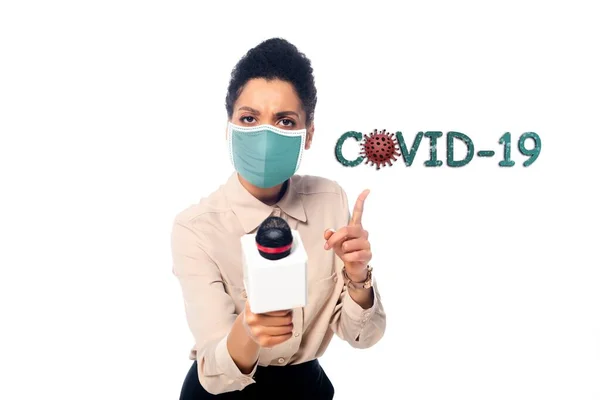 Front view of african american journalist in medical mask with microphone pointing with finger at covid-19 illustration isolated on white — Stock Photo