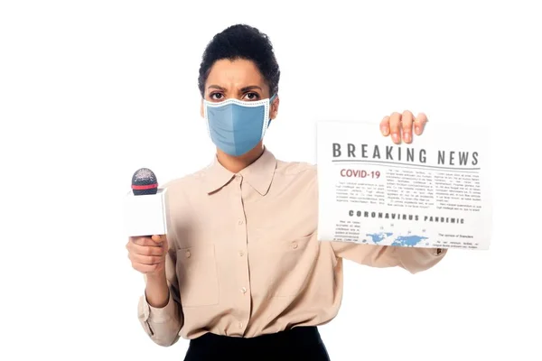 Periodista afroamericano en máscara médica con micrófono mostrando periódico con noticias de última hora covid-19 texto aislado en blanco - foto de stock