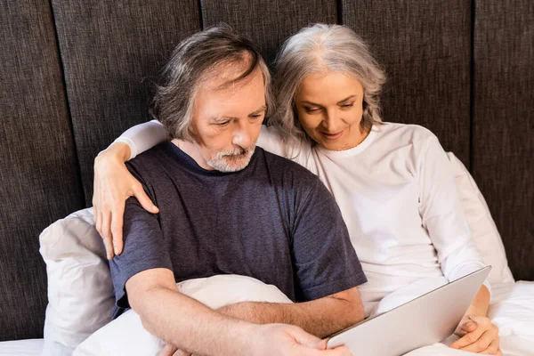 Зрелая пара смотрит на ноутбук в постели дома — стоковое фото