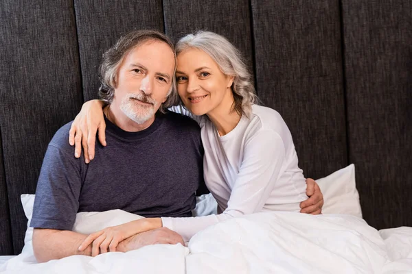 Весёлая зрелая пара обнимается и улыбается в постели — стоковое фото