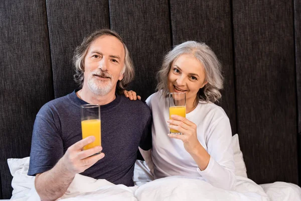 Happy mature couple holding glasses with orange juice in bed — Stock Photo