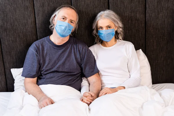 Mature couple in medical masks holding hands in bed — Stock Photo