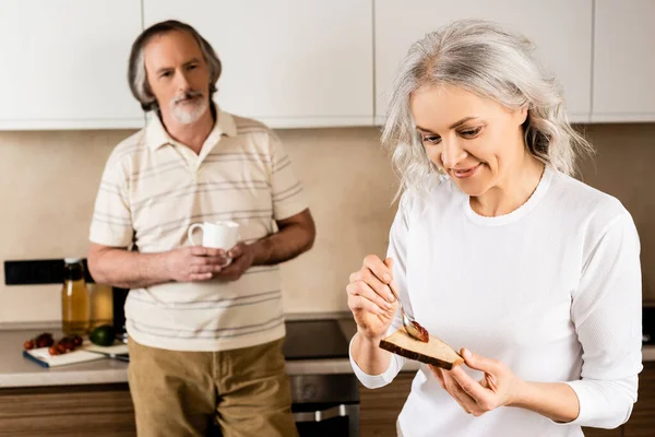 Selective focus of happy mature woman putting jam on toast bread near husband with cup — Stock Photo