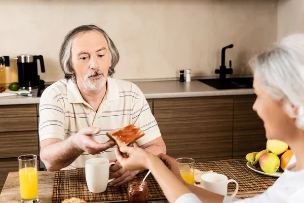 Enfoque selectivo de la mujer madura dando pan tostado dulce con mermelada al marido - foto de stock