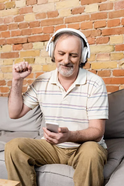 Glücklicher und reifer Mann in drahtlosen Kopfhörern mit Smartphone und Gestik im Wohnzimmer — Stockfoto