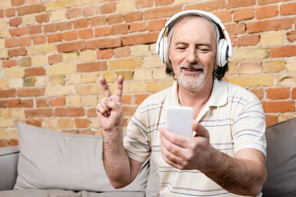 Hombre alegre y maduro en auriculares inalámbricos tomando selfie y mostrando signo de paz en la sala de estar - foto de stock