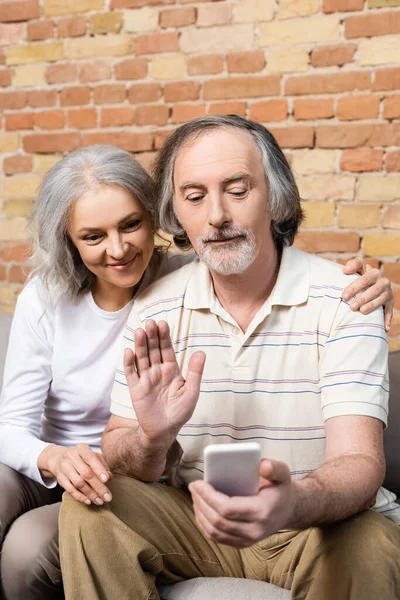 Foco seletivo do homem maduro acenando a mão e olhando para o smartphone com a esposa — Fotografia de Stock