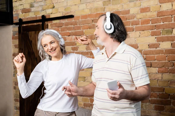 Mujer madura feliz en auriculares inalámbricos bailando cerca de marido con teléfono inteligente - foto de stock