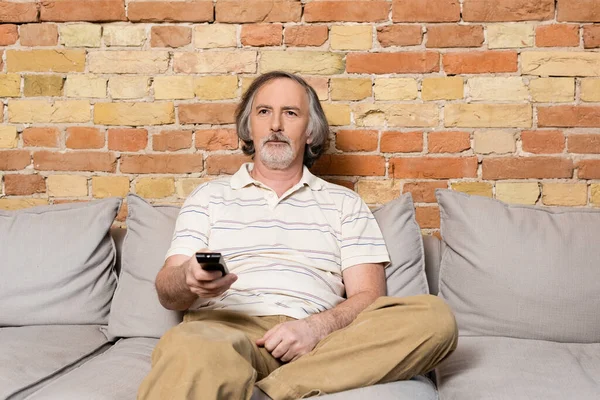 Homme barbu et mature tenant télécommande tout en regardant un film à la maison — Photo de stock