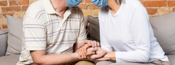 Horizontales Konzept eines reifen Paares in medizinischen Masken, das Händchen hält, während es auf dem Sofa sitzt — Stockfoto