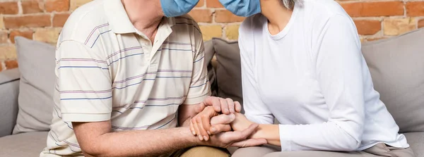 Concepto horizontal de pareja madura en máscaras médicas cogidas de la mano mientras se sienta en el sofá - foto de stock