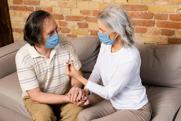 Mature woman in medical mask holding digital thermometer near husband — Stock Photo