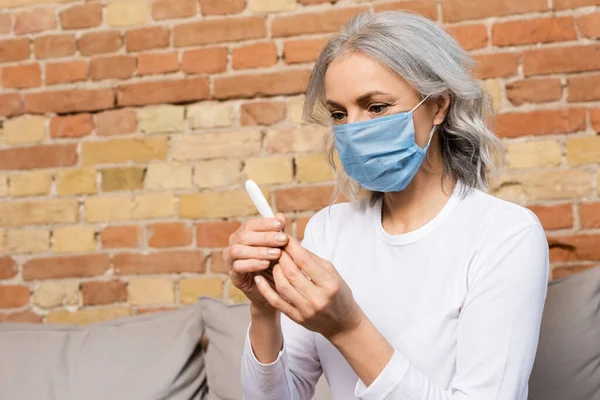 Sick and mature woman in medical mask looking at digital thermometer — Stock Photo