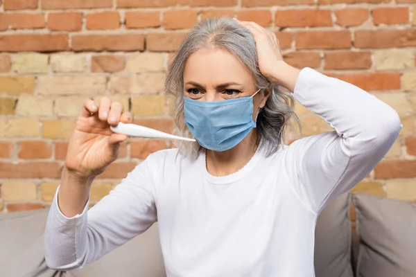 Mature woman in medical mask looking at digital thermometer — Stock Photo
