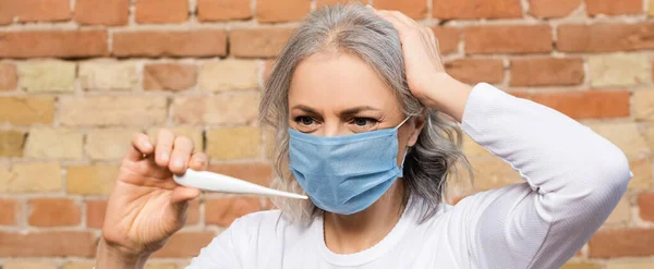 Panoramic orientation of mature woman in medical mask looking at digital thermometer — Stock Photo