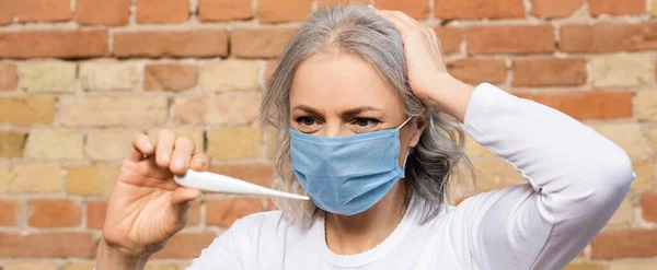 Panoramische Orientierung der reifen Frau in medizinischer Maske beim Betrachten des digitalen Thermometers — Stockfoto