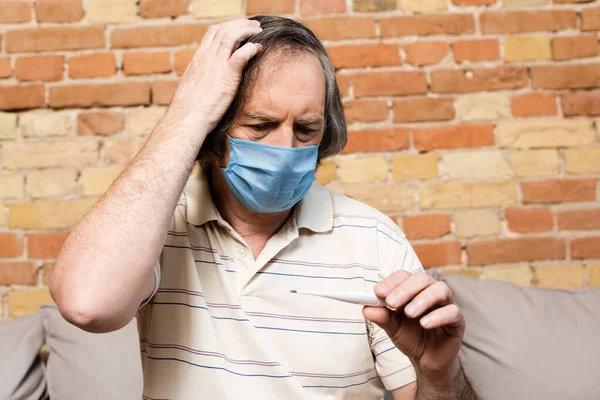 Зрілий чоловік в медичній масці дивиться на цифровий термометр — стокове фото