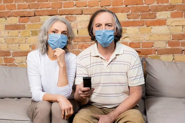 Mature couple in medical masks watching film at home — Stock Photo