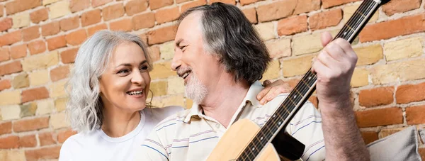 Панорамний знімок бородатого і зрілого чоловіка, який грає на акустичній гітарі біля усміхненої дружини — стокове фото