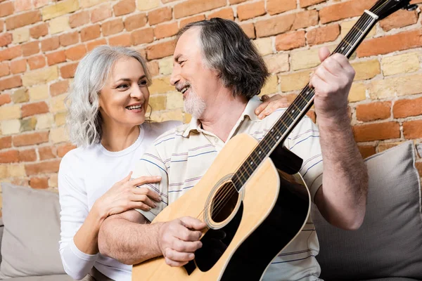 Счастливый и зрелый мужчина играет на акустической гитаре рядом с веселой женой — стоковое фото