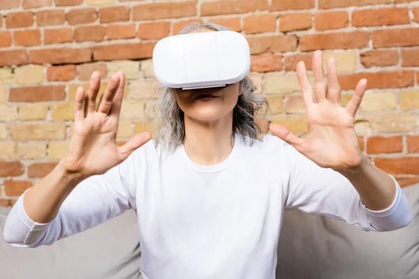 Selective focus of mature woman in virtual reality headset gesturing at home — Stock Photo